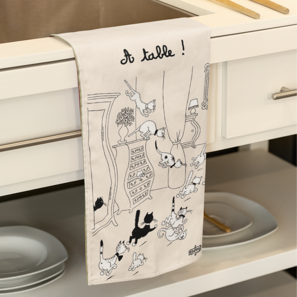 Decorative kitchen towel with black cat illustrations and 'A table!' text hanging over a cabinet.