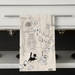 Tea towel with black and white cat illustration hanging on a kitchen cabinet.