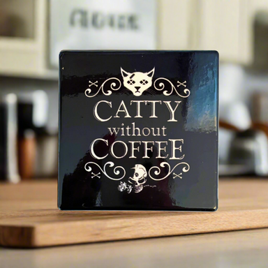 A square-shaped black ceramic coaster with white text and cat designs on it, placed on a wooden surface.