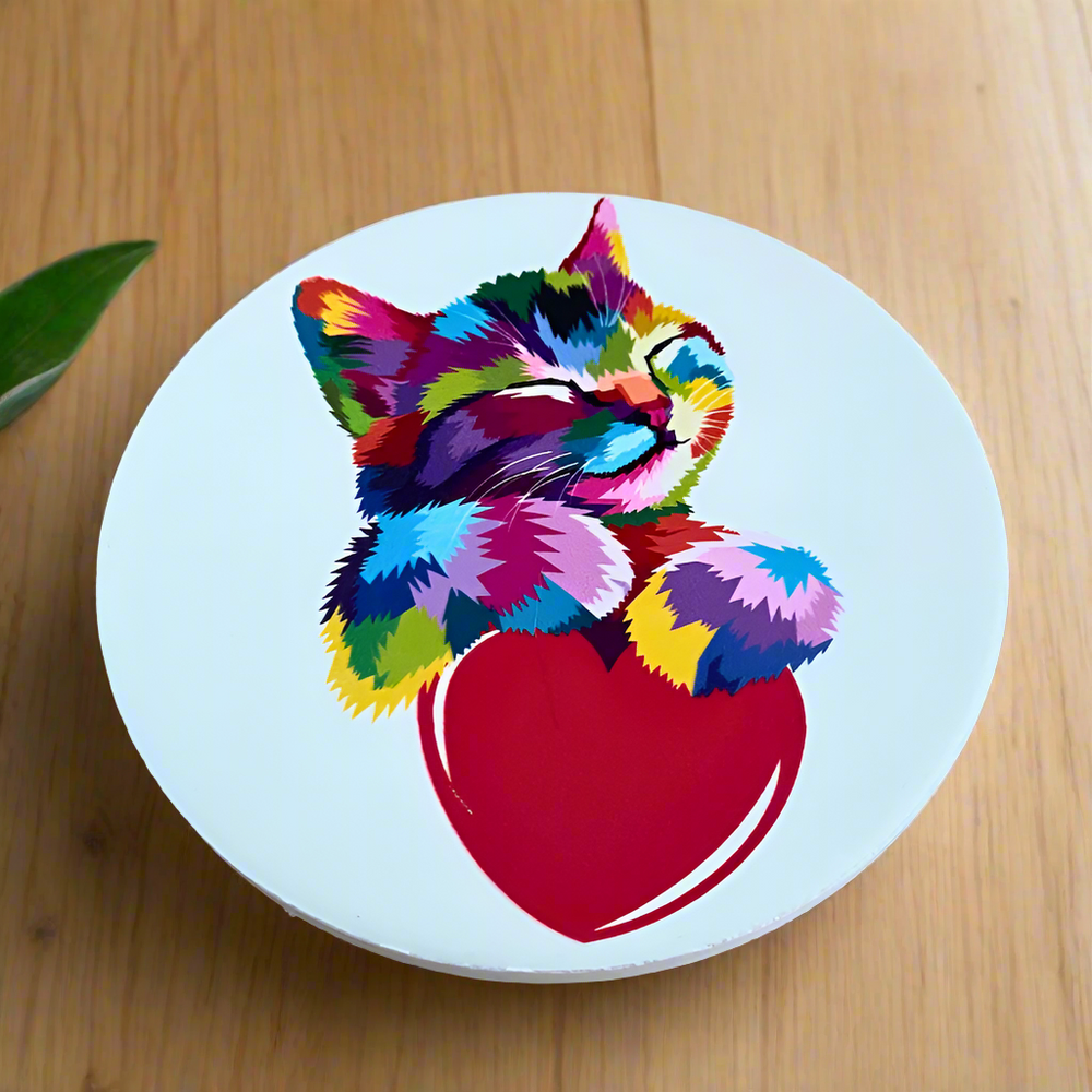 A ceramic round coaster with a rainbow kitten holding a red heart against a white background. The coaster is placed on a wooden surface.