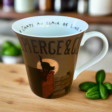 A ceramic mug with a design of a black cat silhouette on a chimney under the moonlight. The mug has white and brown colors, with the artist's signature 'F.
