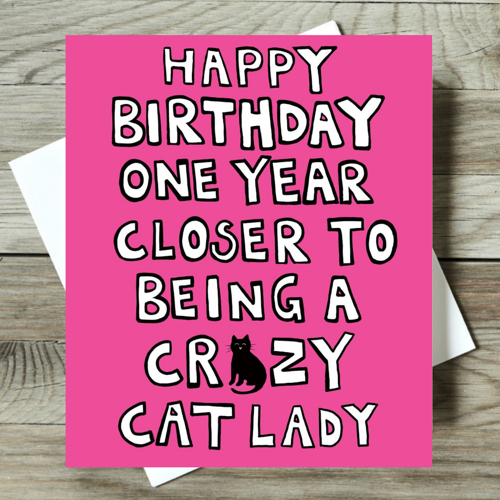 Pink birthday card with text 'Happy Birthday one year closer to being a crazy cat lady' on a wooden surface.