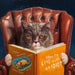 Cat sitting in a leather armchair reading a book titled 'How to Live with an Idiot'.