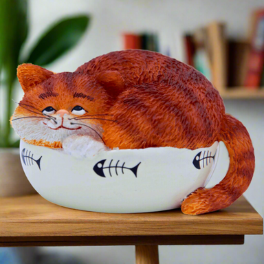A sculpture of a fat ginger cat curled up in a bowl, primarily orange and white in color, made of synthetic resin, positioned on a wooden surface with a plant and books in the background.