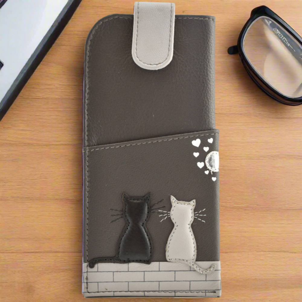 A leather glasses case with a cat design on it, displayed on a wooden table alongside a pair of glasses.