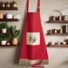 A red apron with a beige border featuring a heart and black cats design, hanging in a kitchen setting with various kitchen items in the background.
