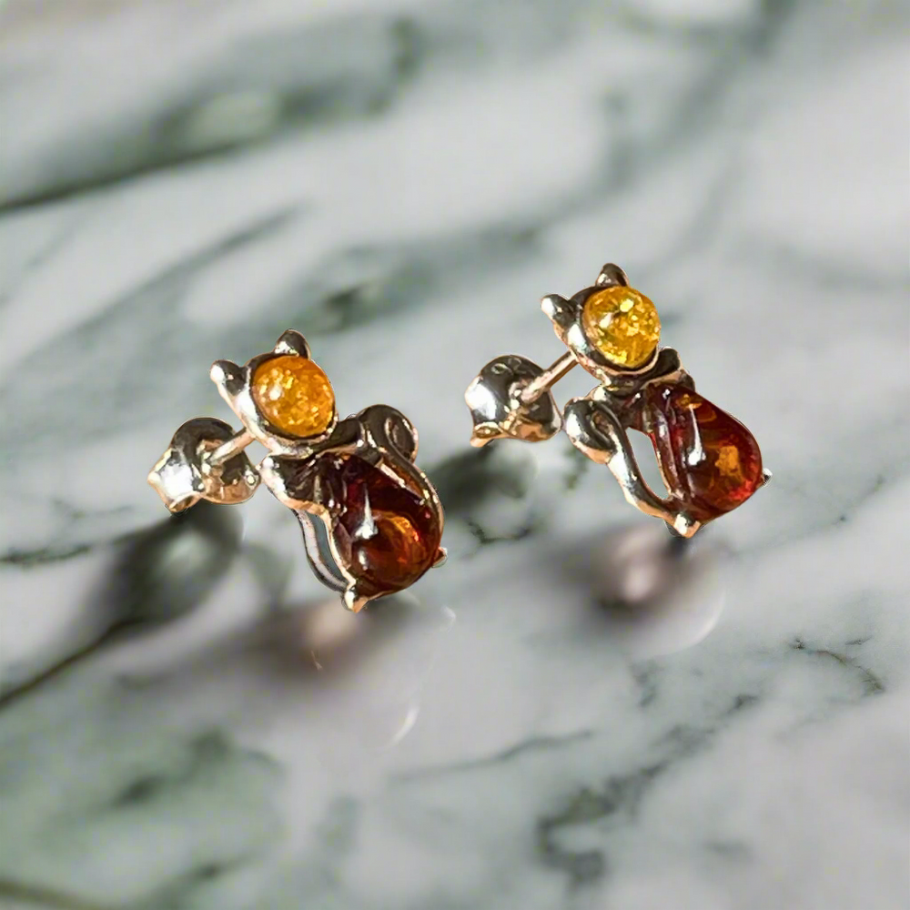 A pair of sterling silver cat-shaped earrings with amber gemstones set in them, displayed on a marble surface.